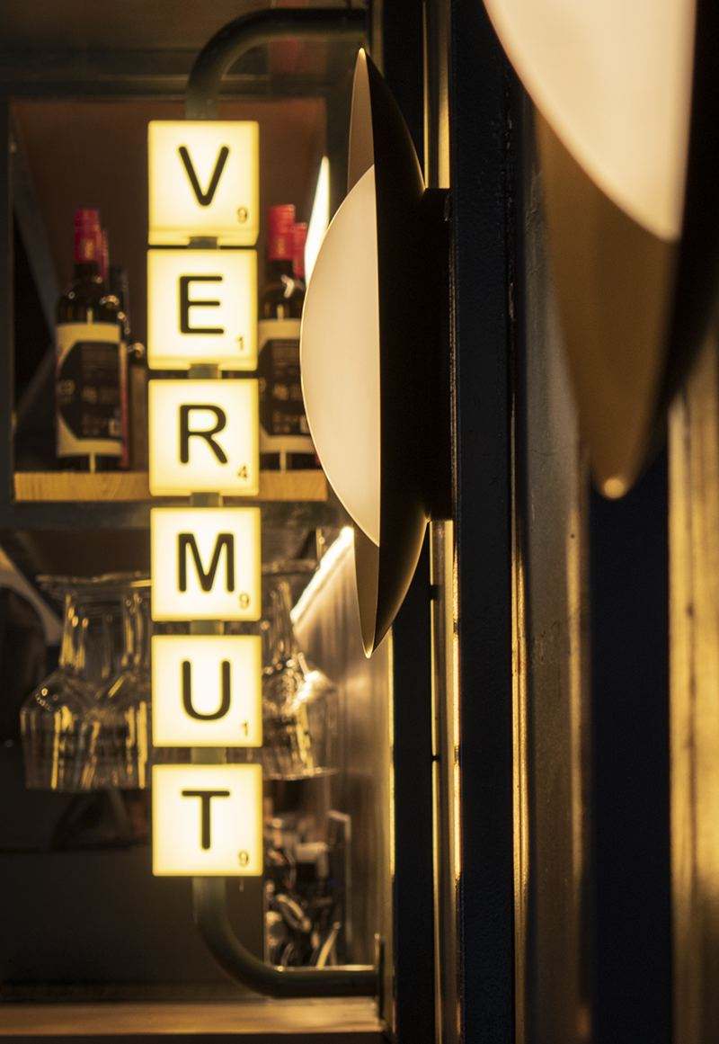 Vertical light box spelling VERMUT in lit letter tiles, next to a round wall lamp and the bottle shelf.