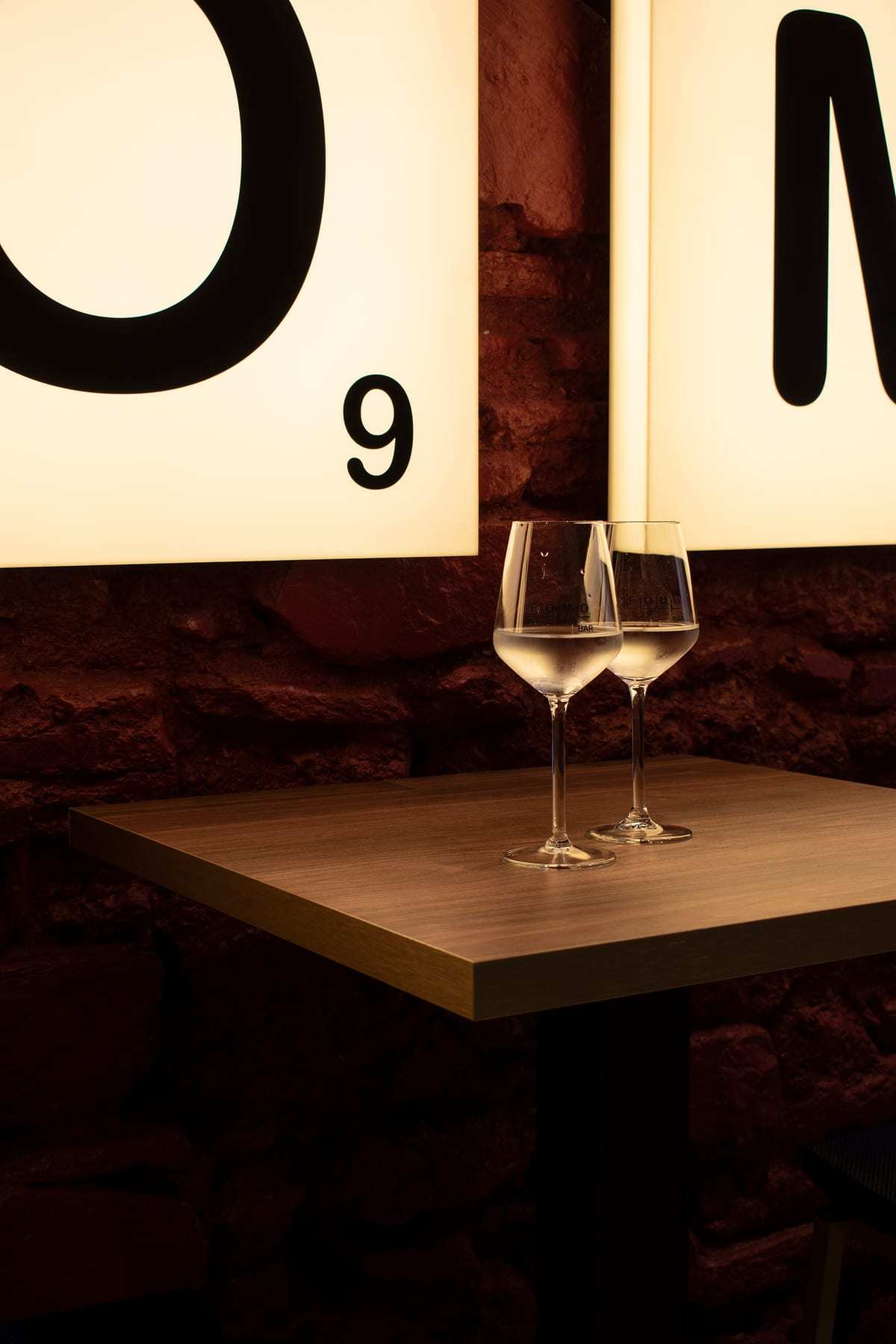 Cajas de luz con las letras O y M sobre la pared de piedra, y una mesa alta con dos copas de vino.
