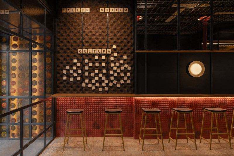 Rincón con la vidriera de posavasos, el corcho lleno de notas bajo las fichas TALLER, MÚSICA y CULTURA, y la barra de azulejo rojo.
