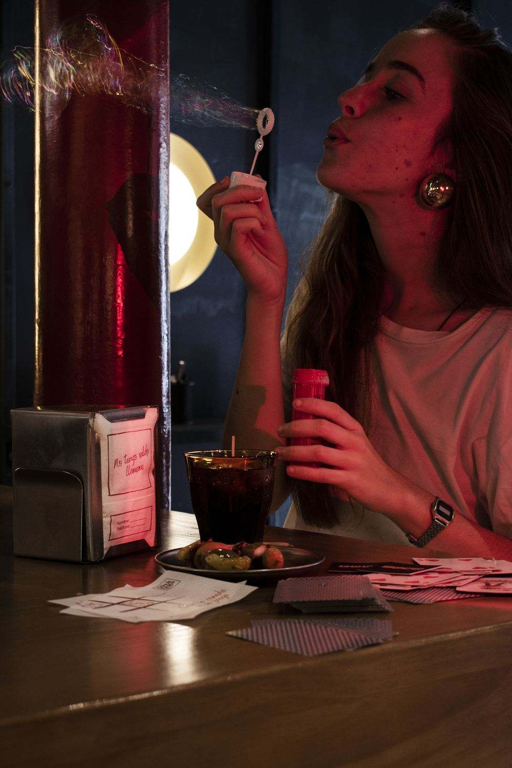 At the bar, a woman blows soap bubbles next to a napkin holder with the “No tengo saldo. Llámame” napkin, a Duralex glass, a small plate of olives, a napkin with noughts and crosses and playing cards.
