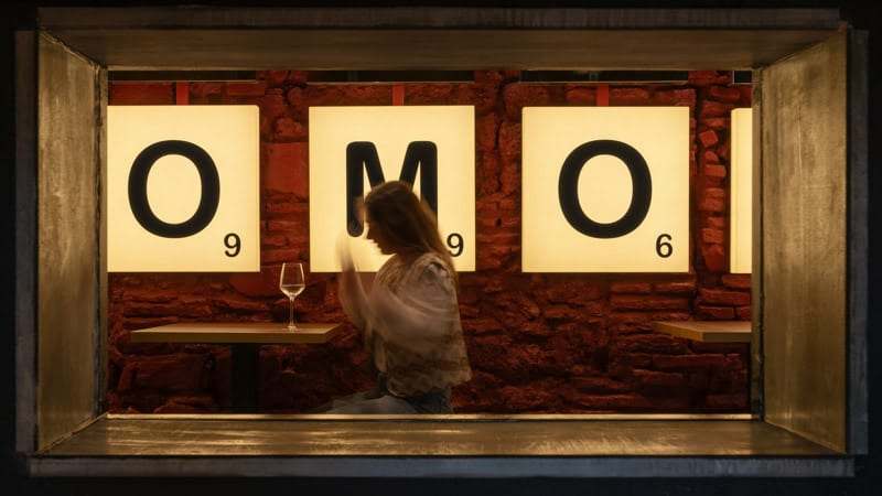 Ventana de FOMO Bar enmarcada en metal con las cajas de luz O, M y O y una clienta tomando vino en la mesa.