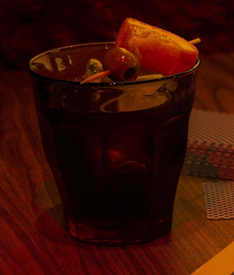 Vermouth in a Duralex glass with ice, orange and a skewered olive, on the wooden table.
