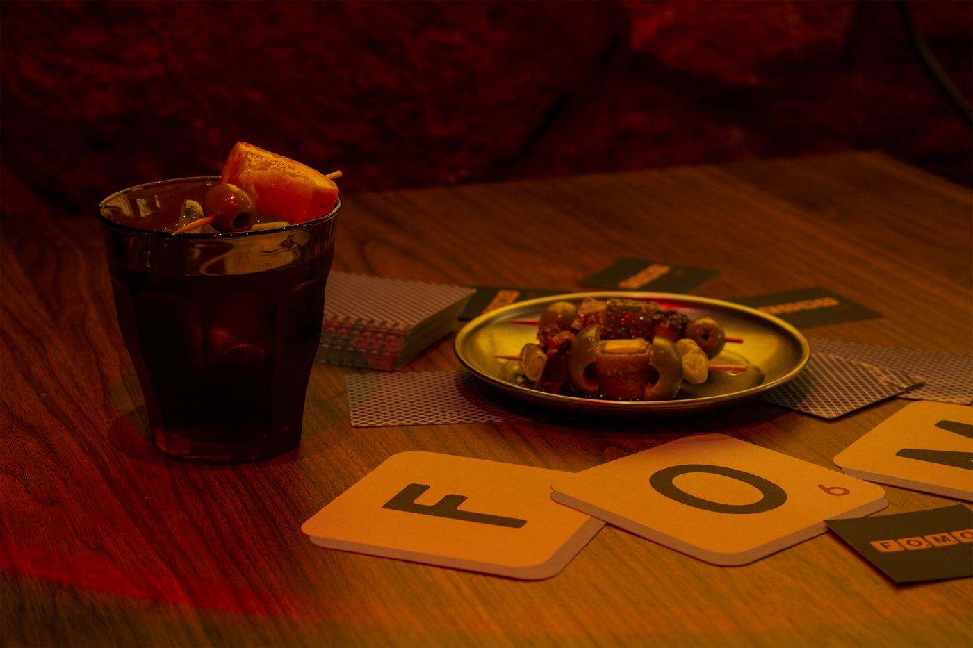Vermouth with orange and a small plate of nibbles on a wooden table, next to coasters shaped like the F and O letter tiles.