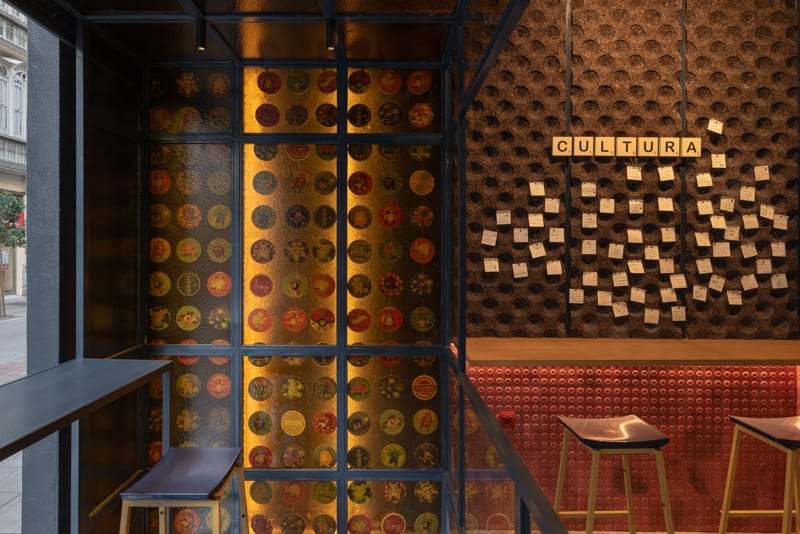 Glass panel of colourful coasters and a cork board full of notes under the CULTURA tiles, with stools by the red-tiled bar.